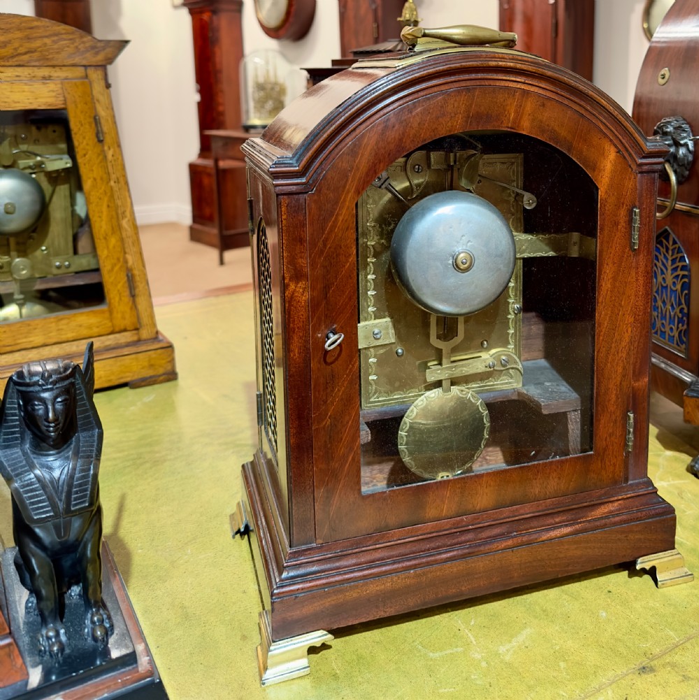 A Regency mahogany bracket clock featuring an arched case, brass carrying handle, circular dial with Roman numerals, and brass bracket feet. View IV