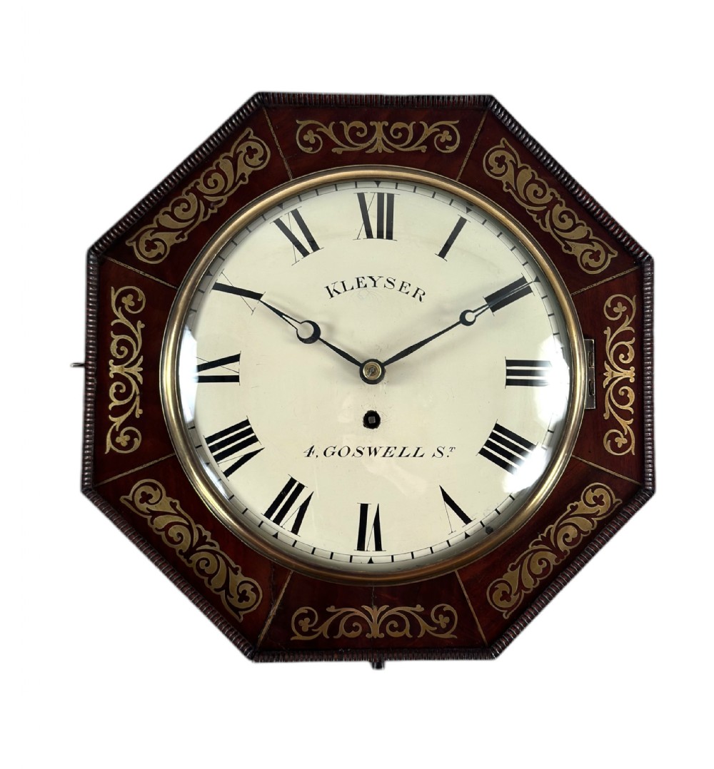 An antique octagonal dark wood wall clock featuring scroll brass inlay surrounding a white dial with Roman numerals.
