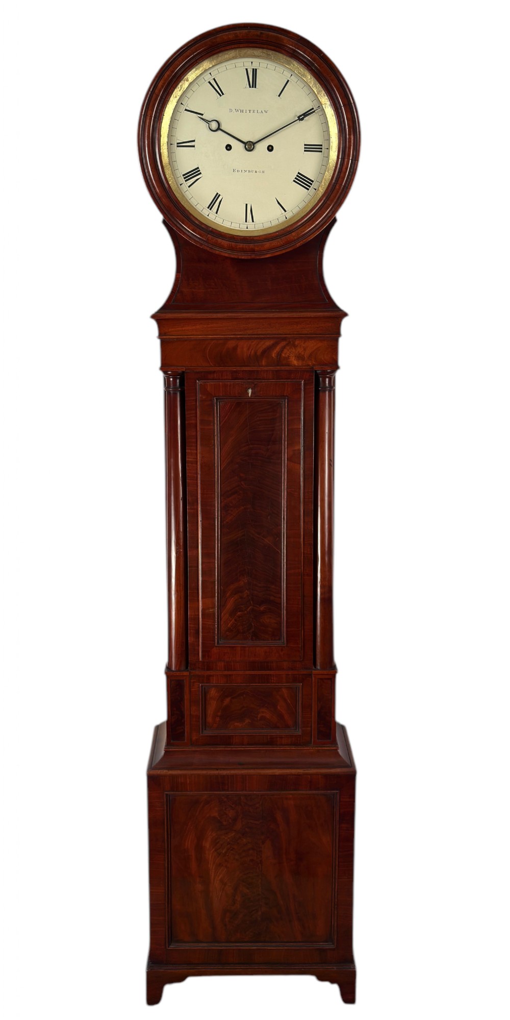 A Scottish Regency flame mahogany longcase clock featuring a circular brass-rimmed dial with Roman numerals and a veneered trunk on bracket feet. View IV