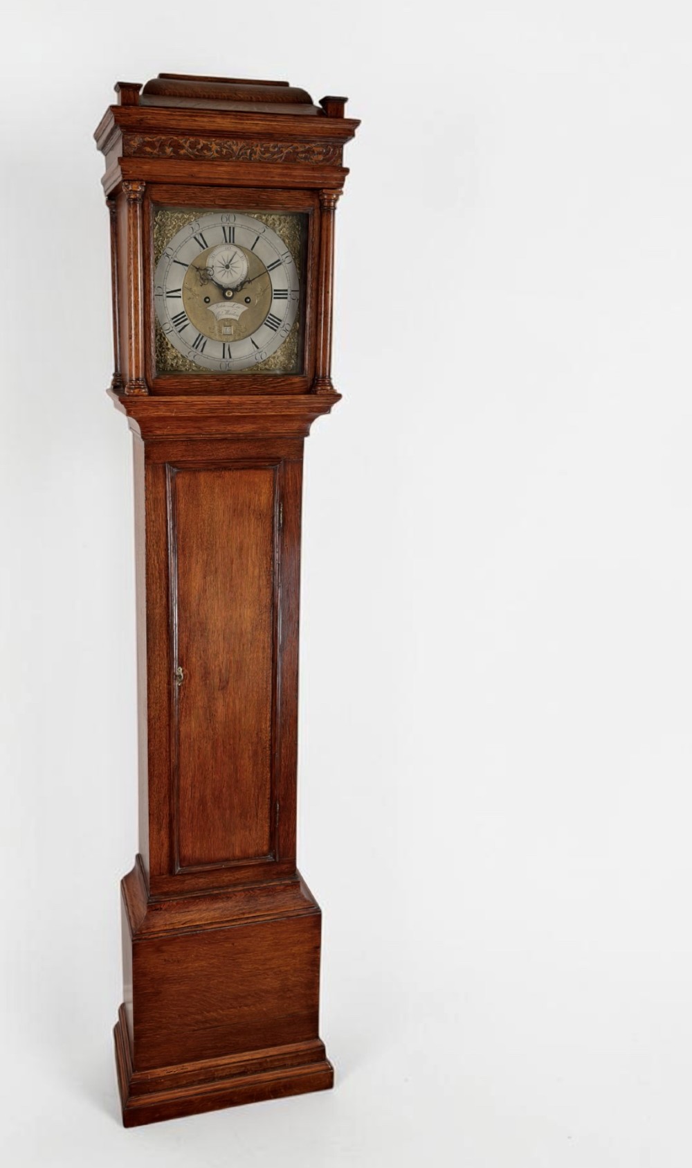 An antique oak longcase clock featuring a carved hood, a brass dial with silvered chapter ring, a narrow trunk door, and a moulded plinth base.