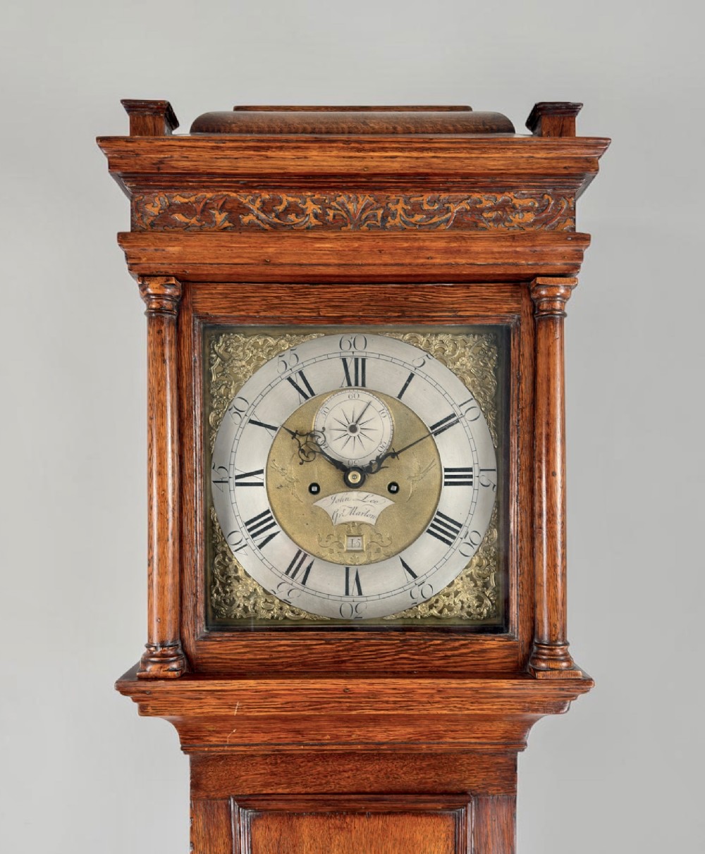 An antique oak longcase clock featuring a carved hood, a brass dial with silvered chapter ring, a narrow trunk door, and a moulded plinth base. View II