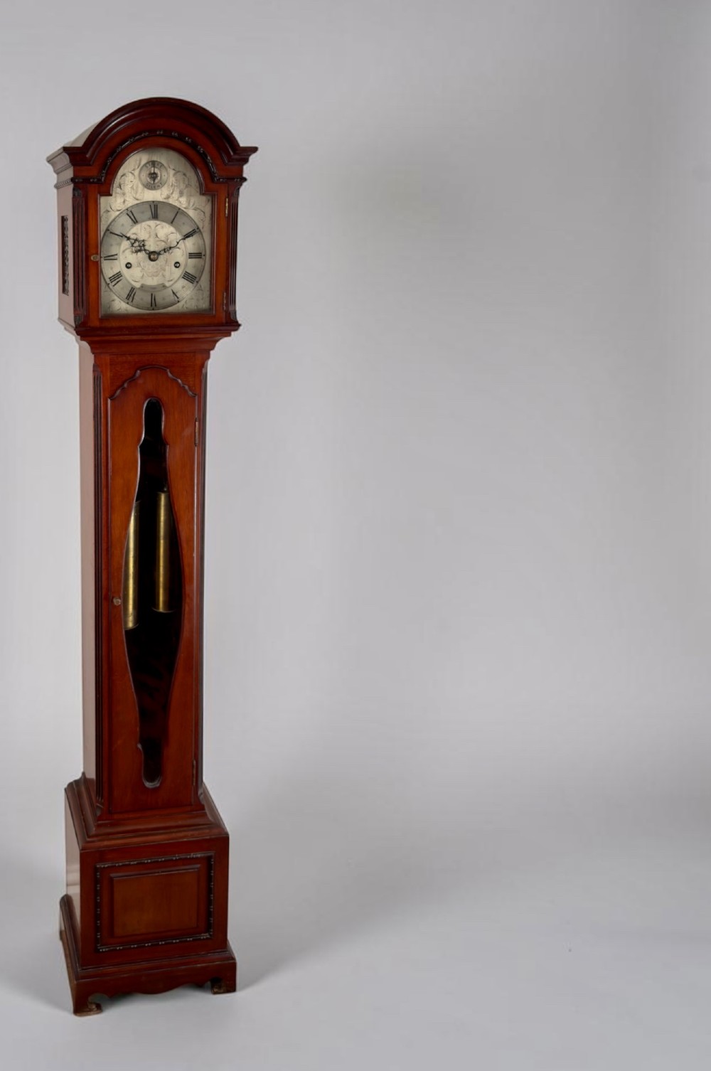 A mahogany grandmother clock featuring an arched hood, a silvered clock dial with Roman numerals, and a glazed trunk displaying brass weights.