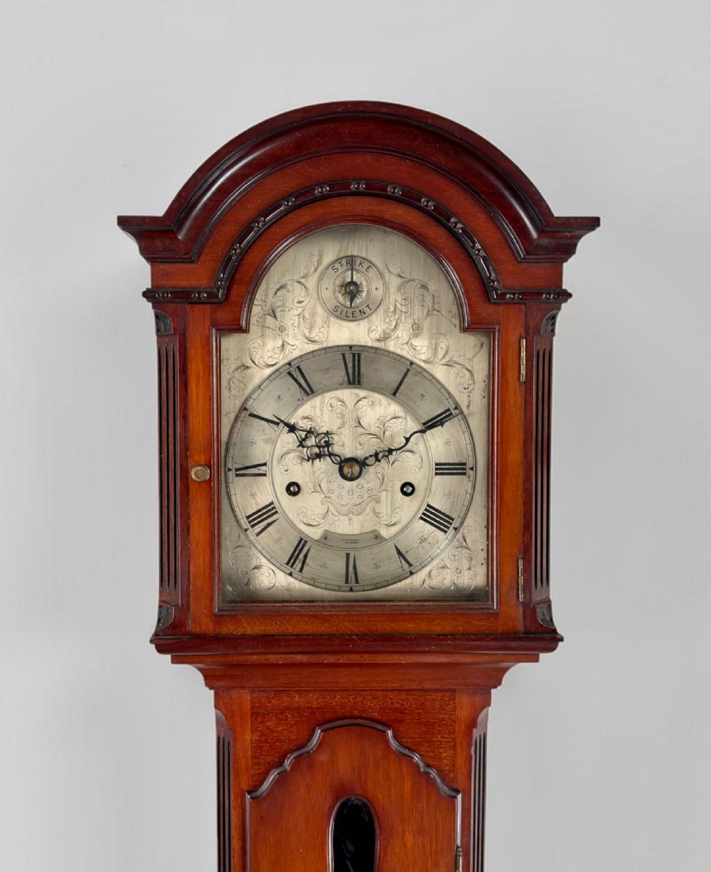 A mahogany grandmother clock featuring an arched hood, a silvered clock dial with Roman numerals, and a glazed trunk displaying brass weights. View III
