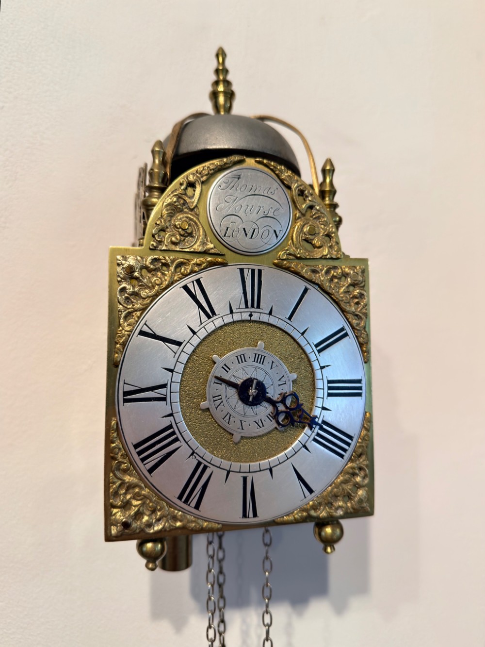 An antique miniature brass hook and spike wall clock with an engraved silvered chapter ring, ornate spandrels, and a top bell assembly.