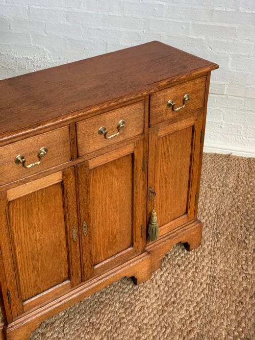 fig 2. A golden oak sideboard featuring four upper drawers, four panelled cupboard doors with brass handles, keyhole escutcheons with tassels, and bracket feet.