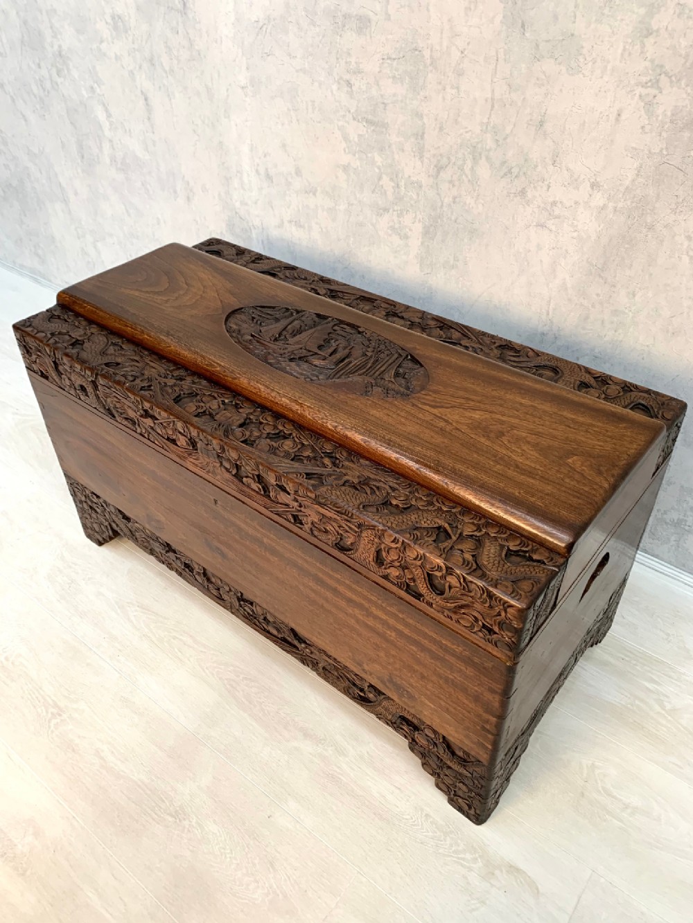 An elevated, high-angle view of a dark brown carved wooden chest featuring detailed low-relief panels depicting traditional motifs and a hinged lid.