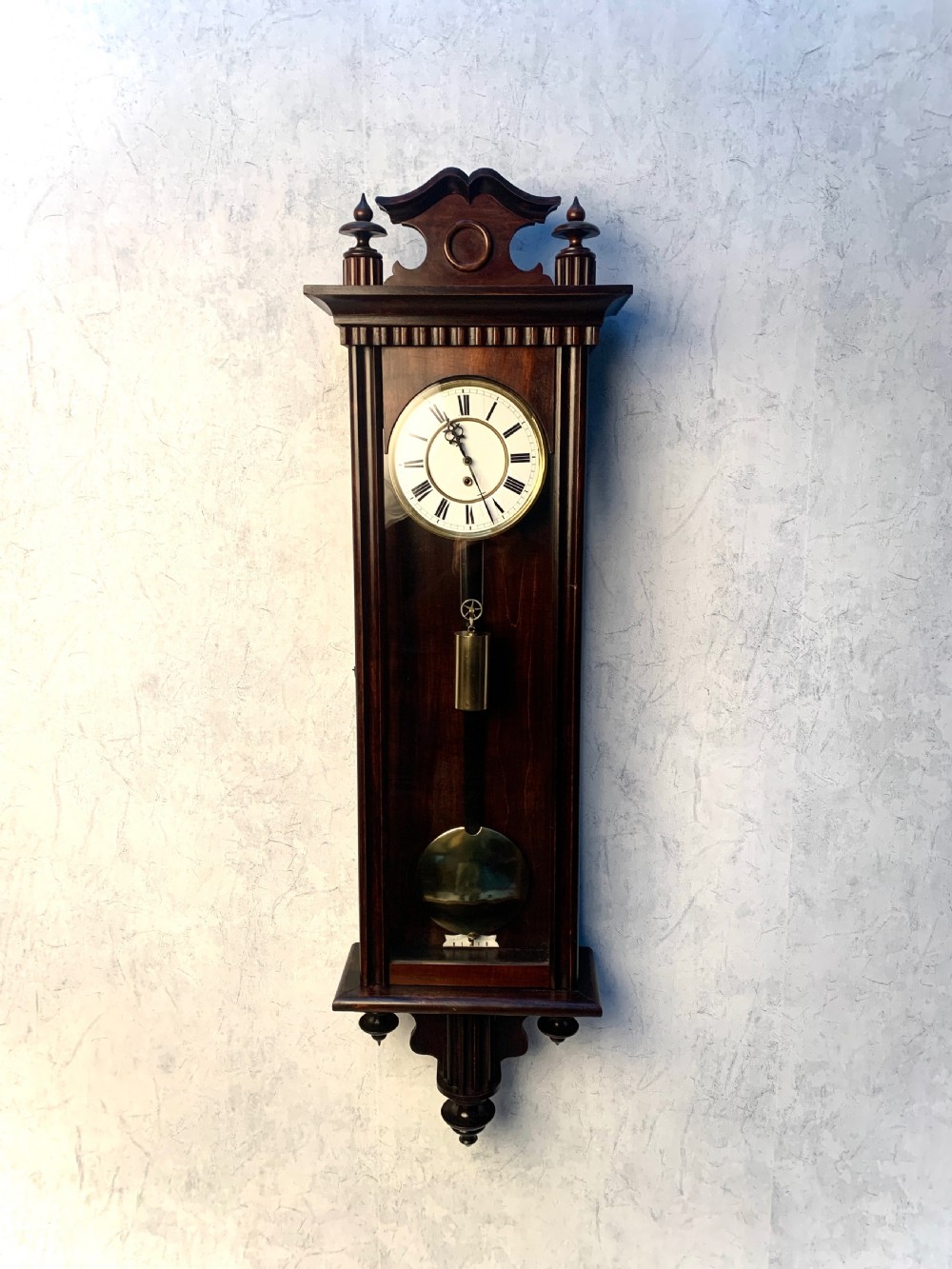 An antique Biedermeier Vienna regulator wall clock featuring a dark mahogany glazed case, carved cresting, white enamel dial with Roman numerals, and a visible brass pendulum.