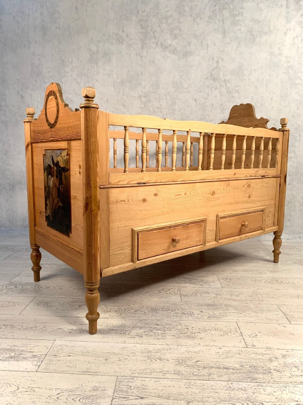 An antique Victorian pine childs bed featuring turned side spindles, two base drawers, corner finials, and a painted pictorial panel.