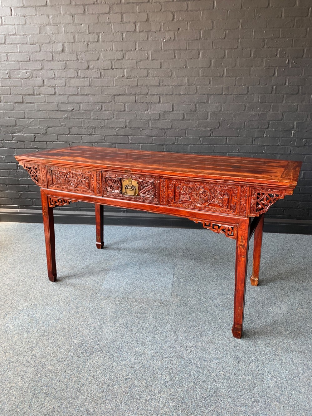 A long Chinese red lacquered altar table featuring detailed hand-carved front panels, central brass hardware, and decorative spandrels.