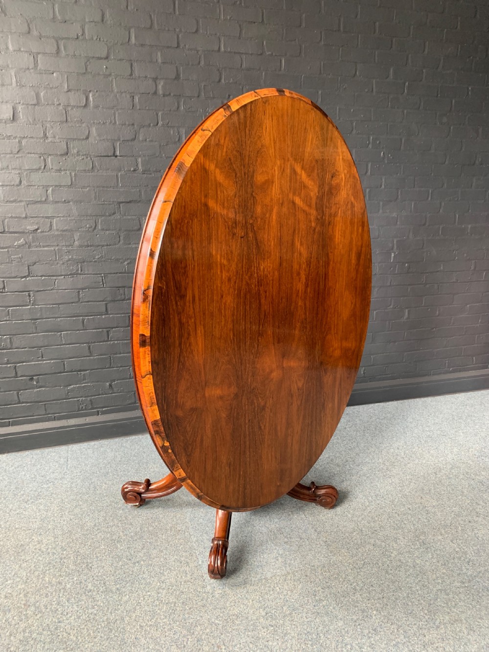 A Victorian rosewood oval tilt-top centre table with a highly figured grain top tilted vertically on a carved pedestal base with scroll feet.