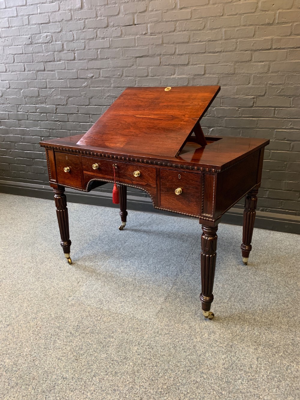 An antique Regency rosewood desk featuring an elevated central writing slope, three drawers, and reeded legs with brass castors.