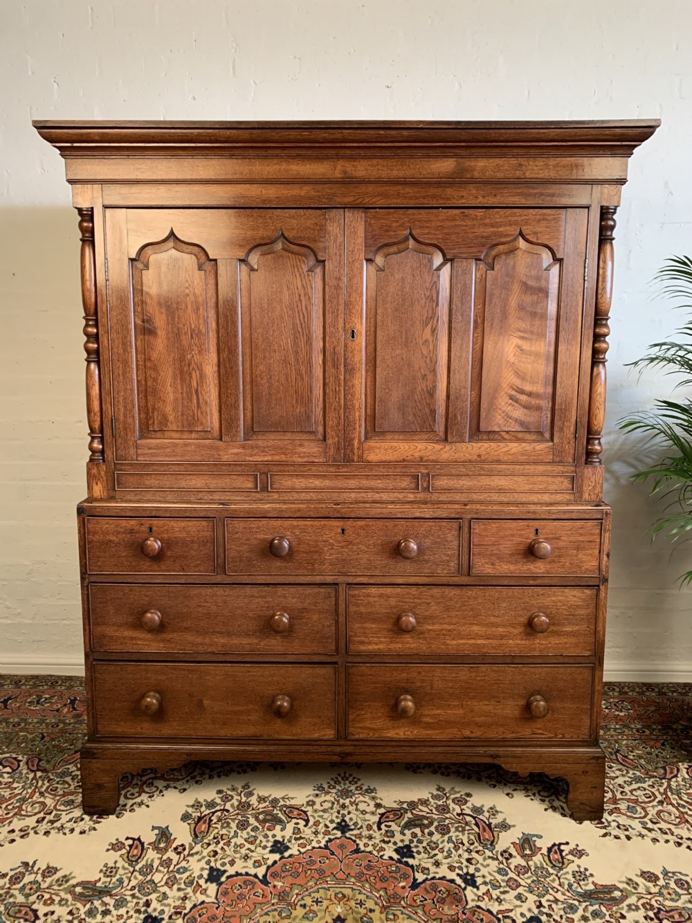 A warm brown oak linen press featuring an upper cupboard with double arched panel doors and turned columns over a multi-drawer base.
