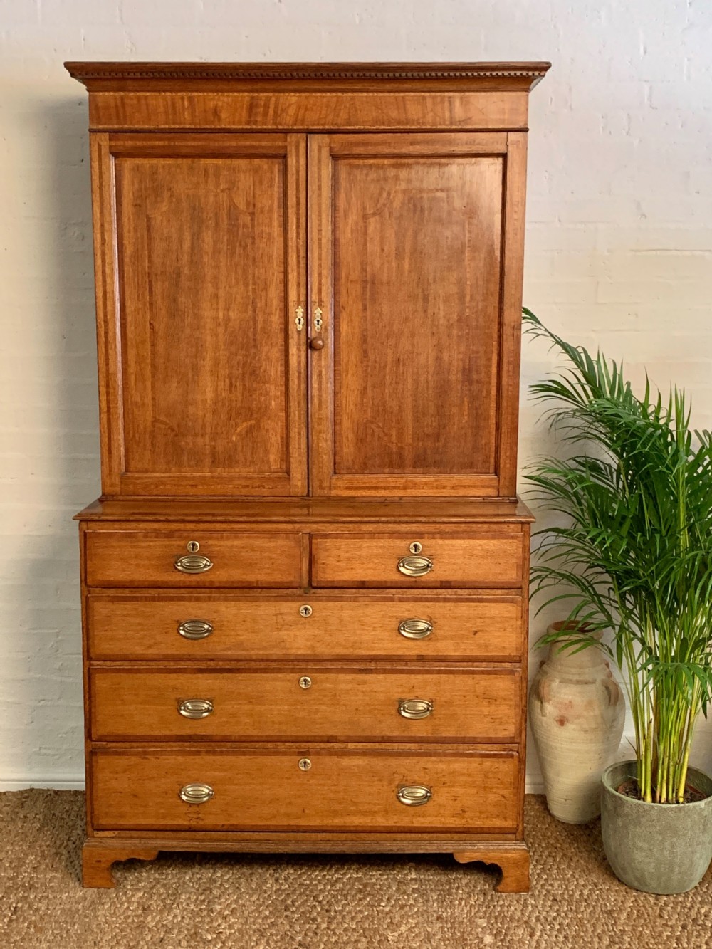 A two-stage Georgian oak press cupboard featuring a double-door upper cabinet mounted over a chest of drawers with oval brass handles.