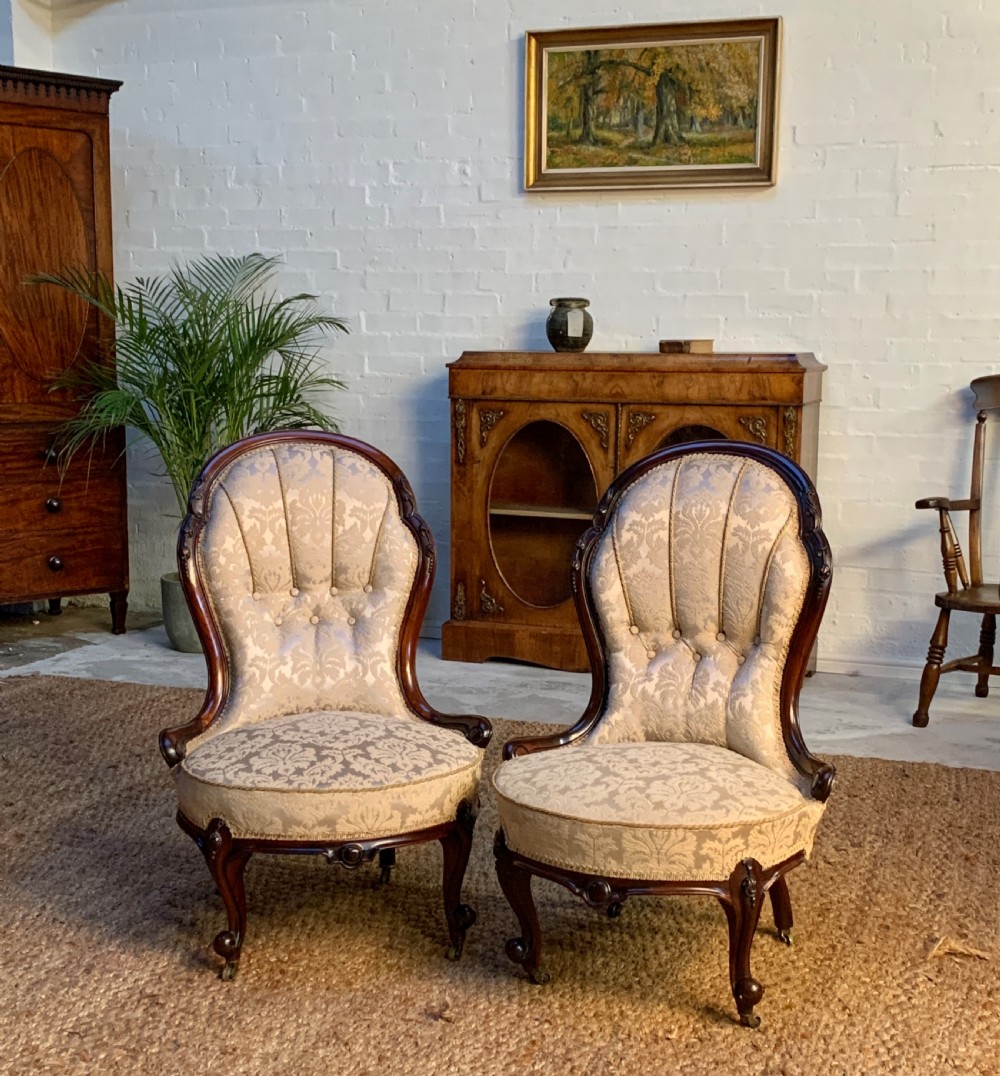 A pair of low-seated Victorian salon chairs featuring dark carved wood frames, fluted shell-back cream damask upholstery, and cabriole legs on brass castors.