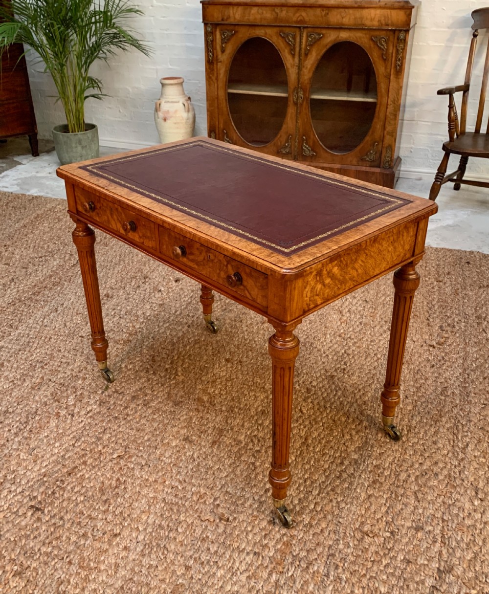 A Victorian burr walnut writing desk featuring a gilt-tooled red leather inset surface, frieze drawers with wooden turned knobs, and reeded legs terminating in brass castors.