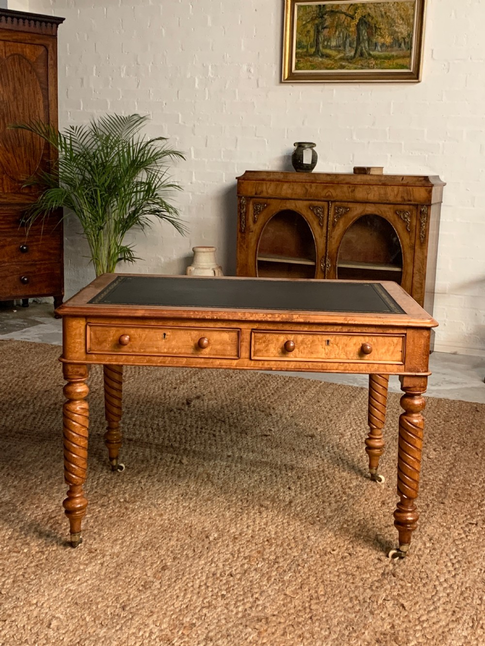 A Victorian golden wood writing desk featuring a dark leather-inset top, two frieze drawers with wooden knob handles, and turned spiral legs supported on brass castors.