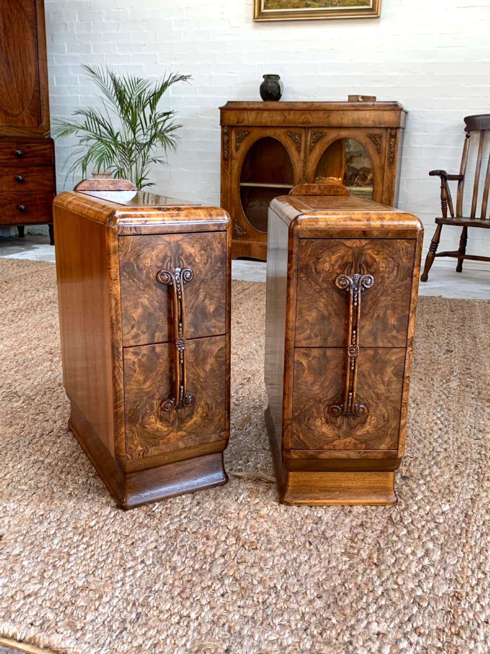 A pair of narrow Art Deco burr walnut bedside cabinets featuring curved upper edges, vertical scroll handles, and plinth bases.