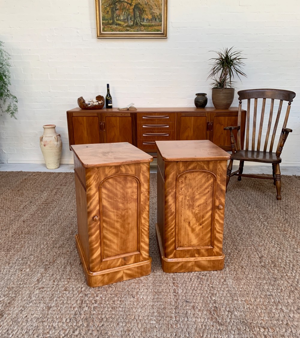 A pair of Victorian satinwood bedside cabinets featuring arched panel doors, turned wooden handles, and plinth bases.