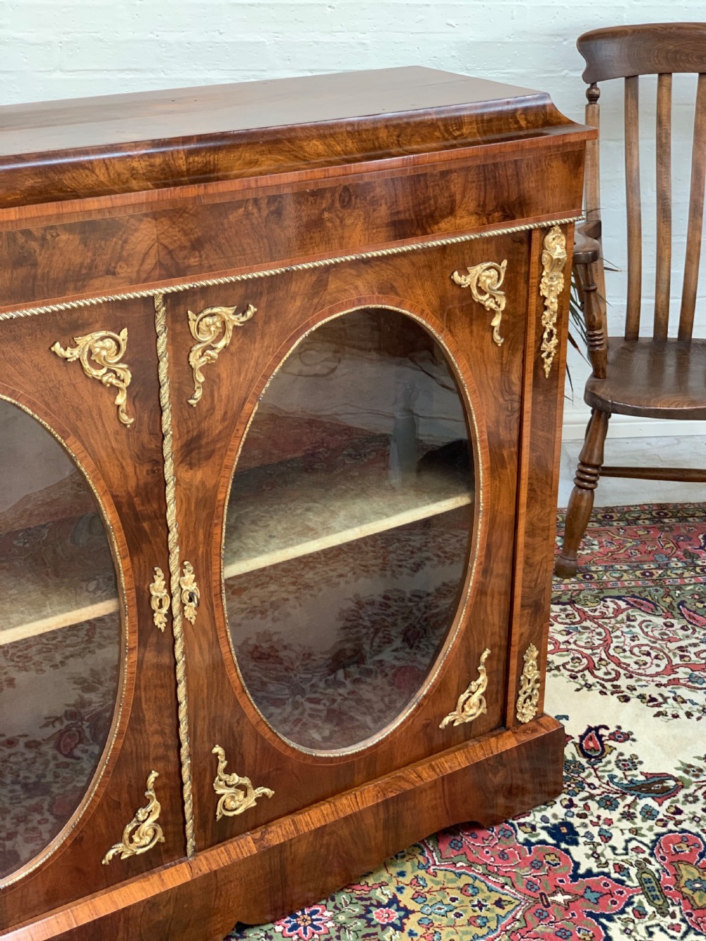 A Victorian double-door pier cabinet featuring figured walnut veneers, oval glass door panels, and decorative brass mounts. View VI