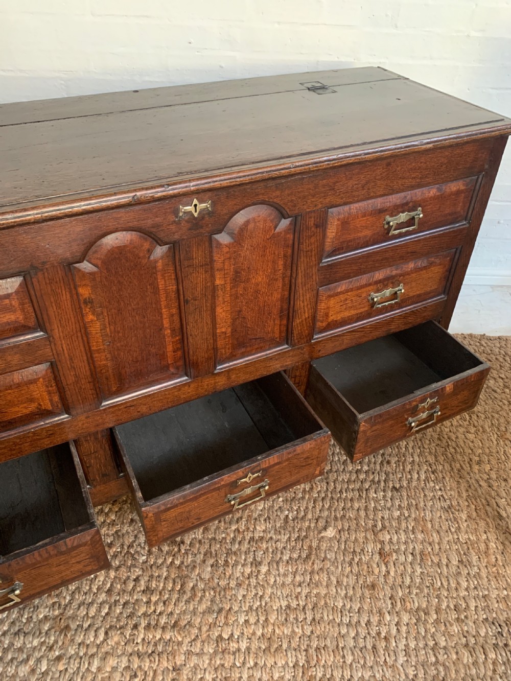 A dark oak mule chest featuring a hinged top, raised arched panels, multiple lower drawers, and brass batwing handles. View XII