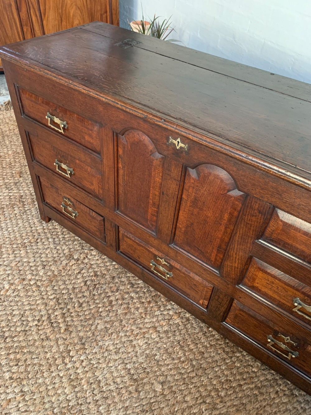 A dark oak mule chest featuring a hinged top, raised arched panels, multiple lower drawers, and brass batwing handles. View XIII
