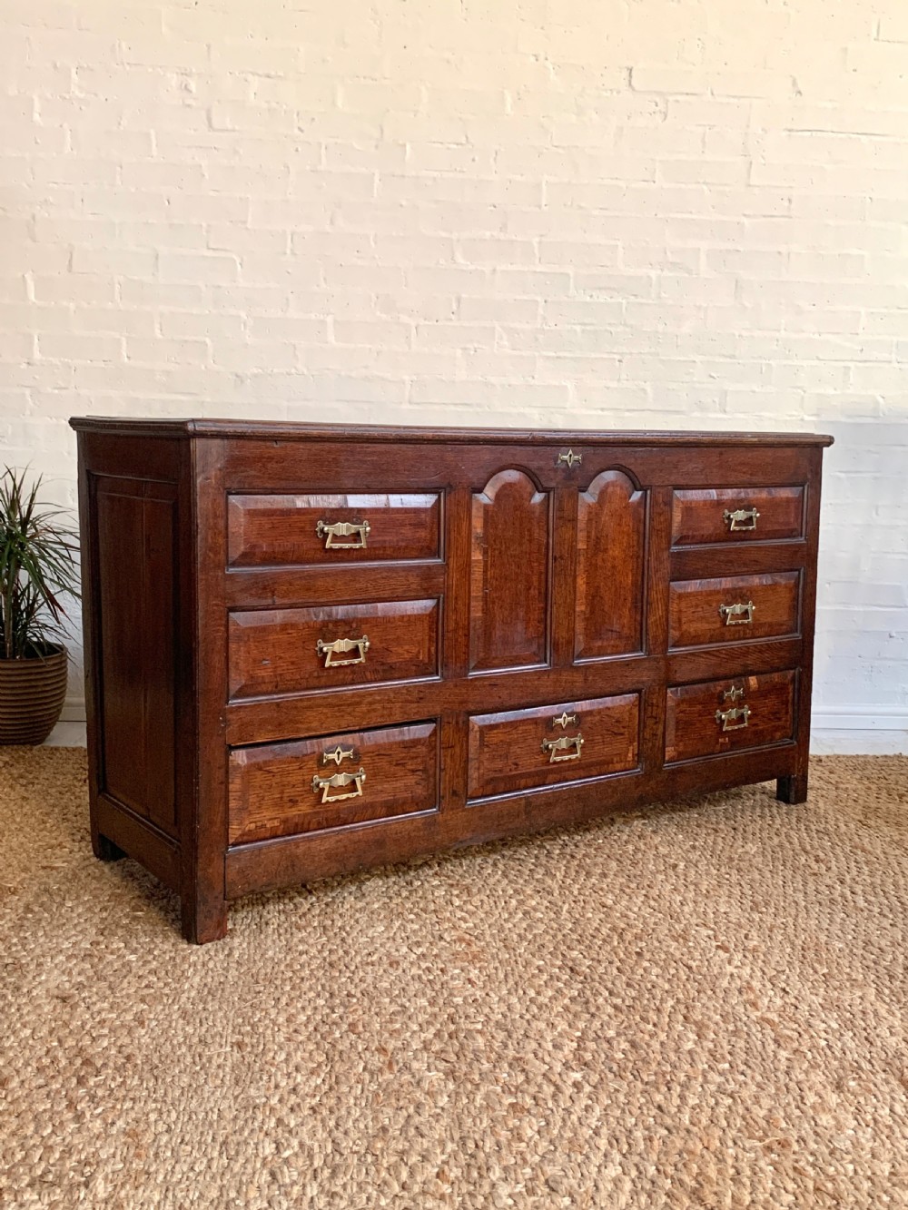 A dark oak mule chest featuring a hinged top, raised arched panels, multiple lower drawers, and brass batwing handles. View XVI