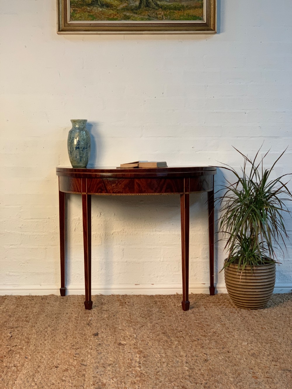 A Georgian style mahogany demi-lune card table with an inlaid apron and tapered square legs standing against a white wall.