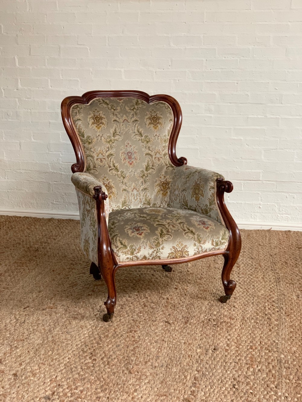 A Victorian mahogany gentlemans armchair upholstered in sage green patterned damask fabric with carved scroll arms and cabriole legs on brass castors.