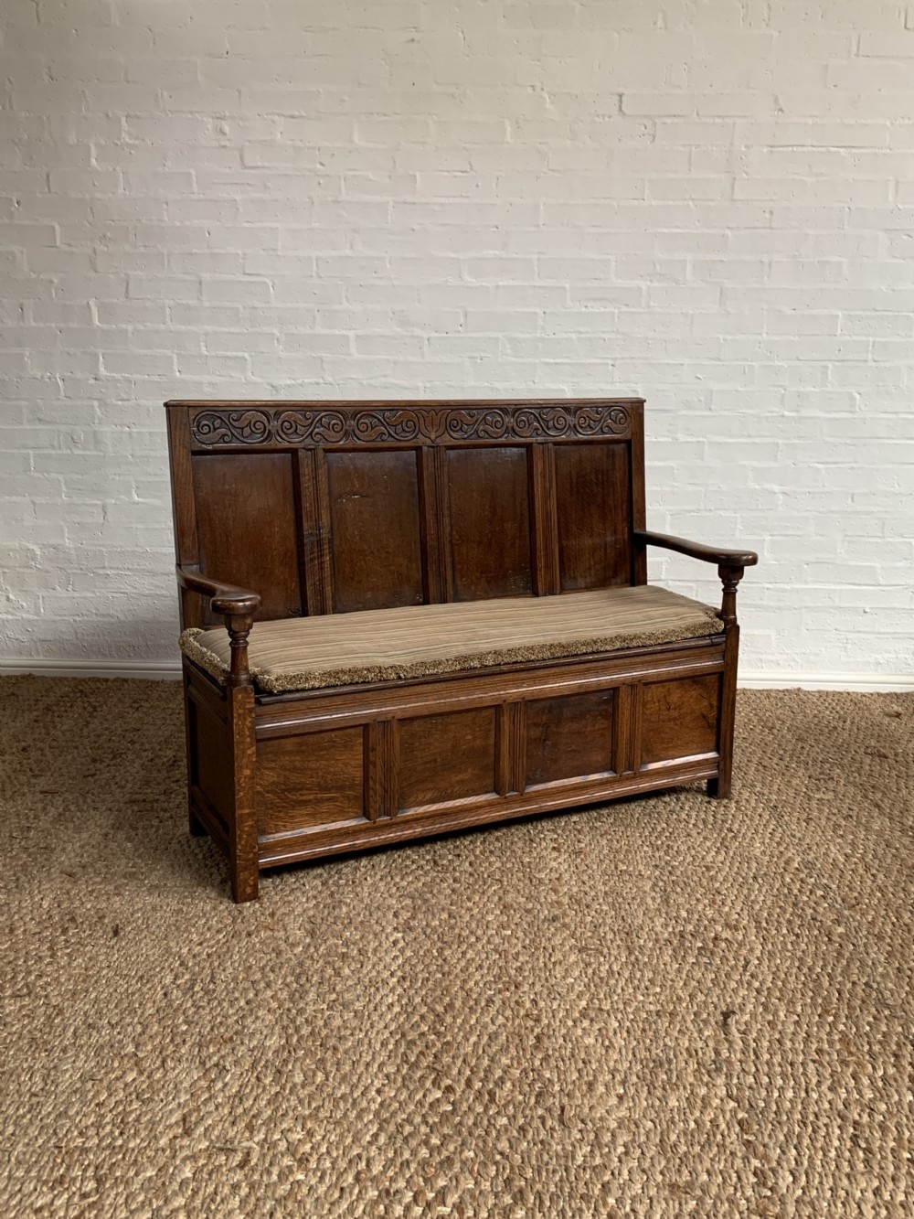 A dark oak hall bench featuring a carved upper frieze, panelled back, turned arm supports, and a fabric-upholstered seat over a panelled storage base.