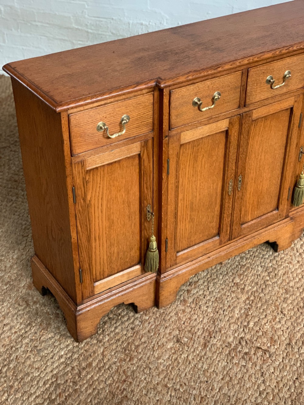A golden oak sideboard featuring four upper drawers, four panelled cupboard doors with brass handles, keyhole escutcheons with tassels, and bracket feet. View II
