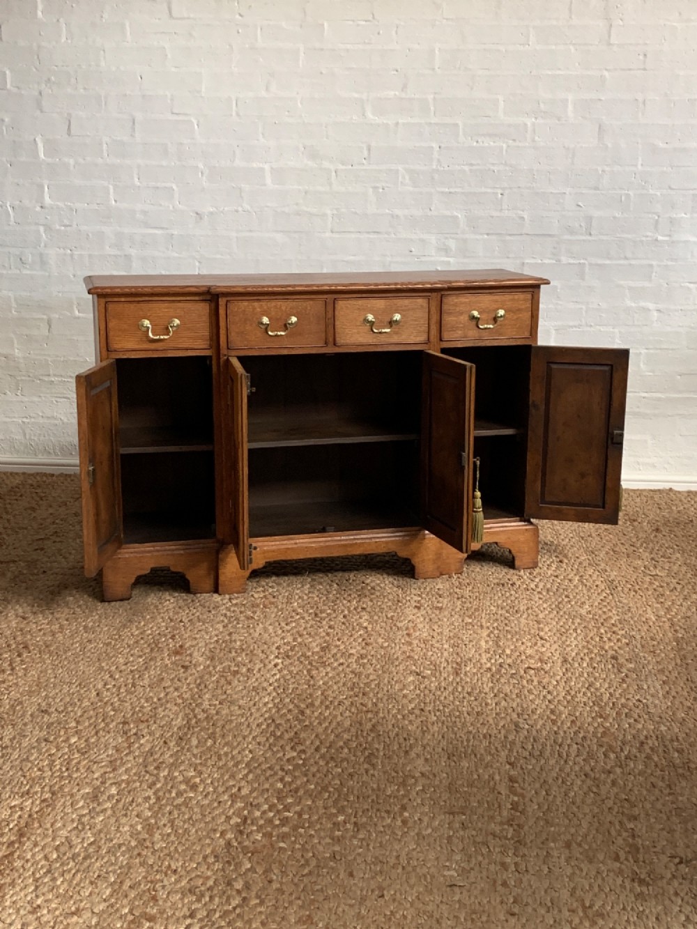A golden oak sideboard featuring four upper drawers, four panelled cupboard doors with brass handles, keyhole escutcheons with tassels, and bracket feet. View V