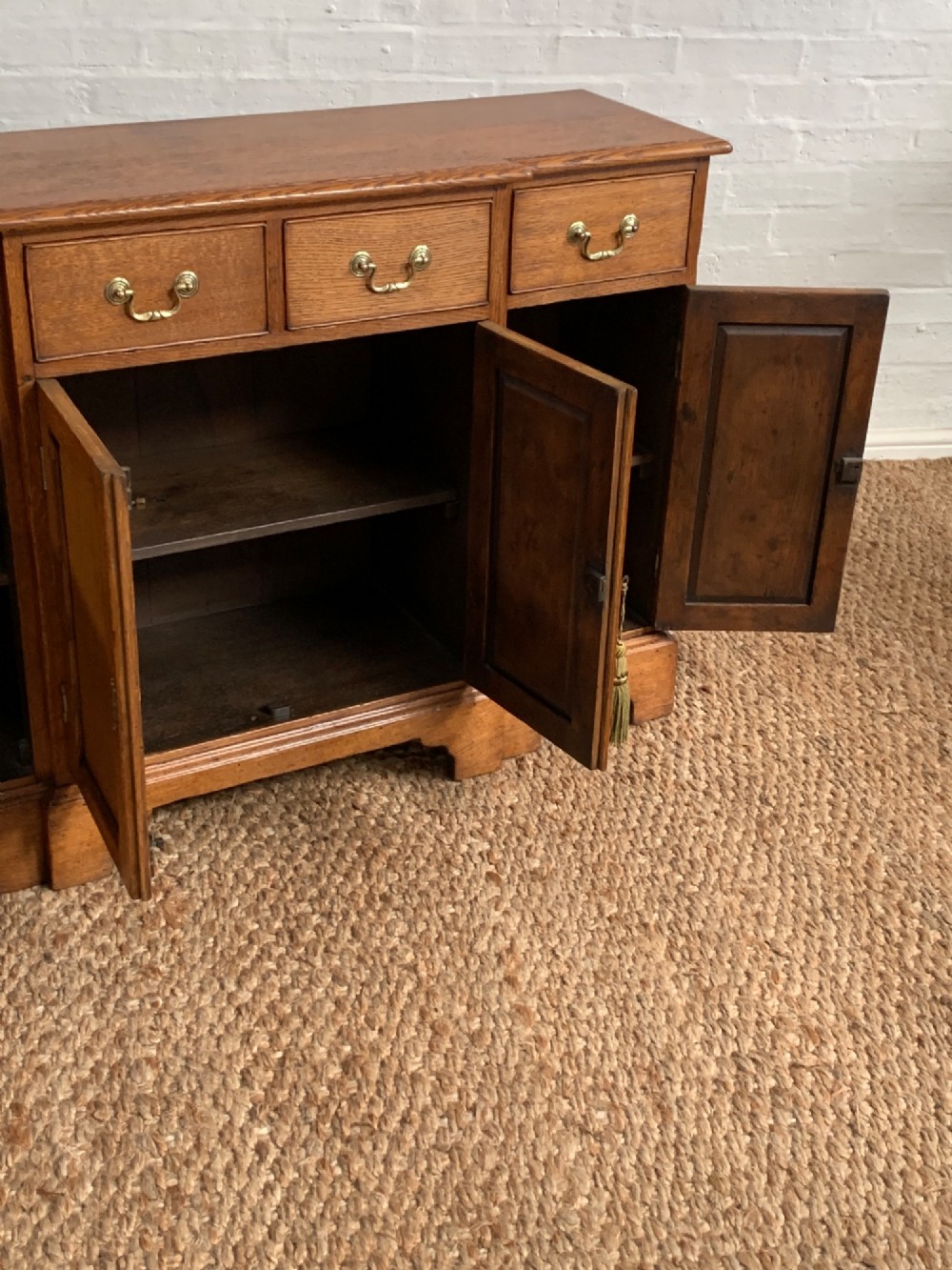 A golden oak sideboard featuring four upper drawers, four panelled cupboard doors with brass handles, keyhole escutcheons with tassels, and bracket feet. View VII