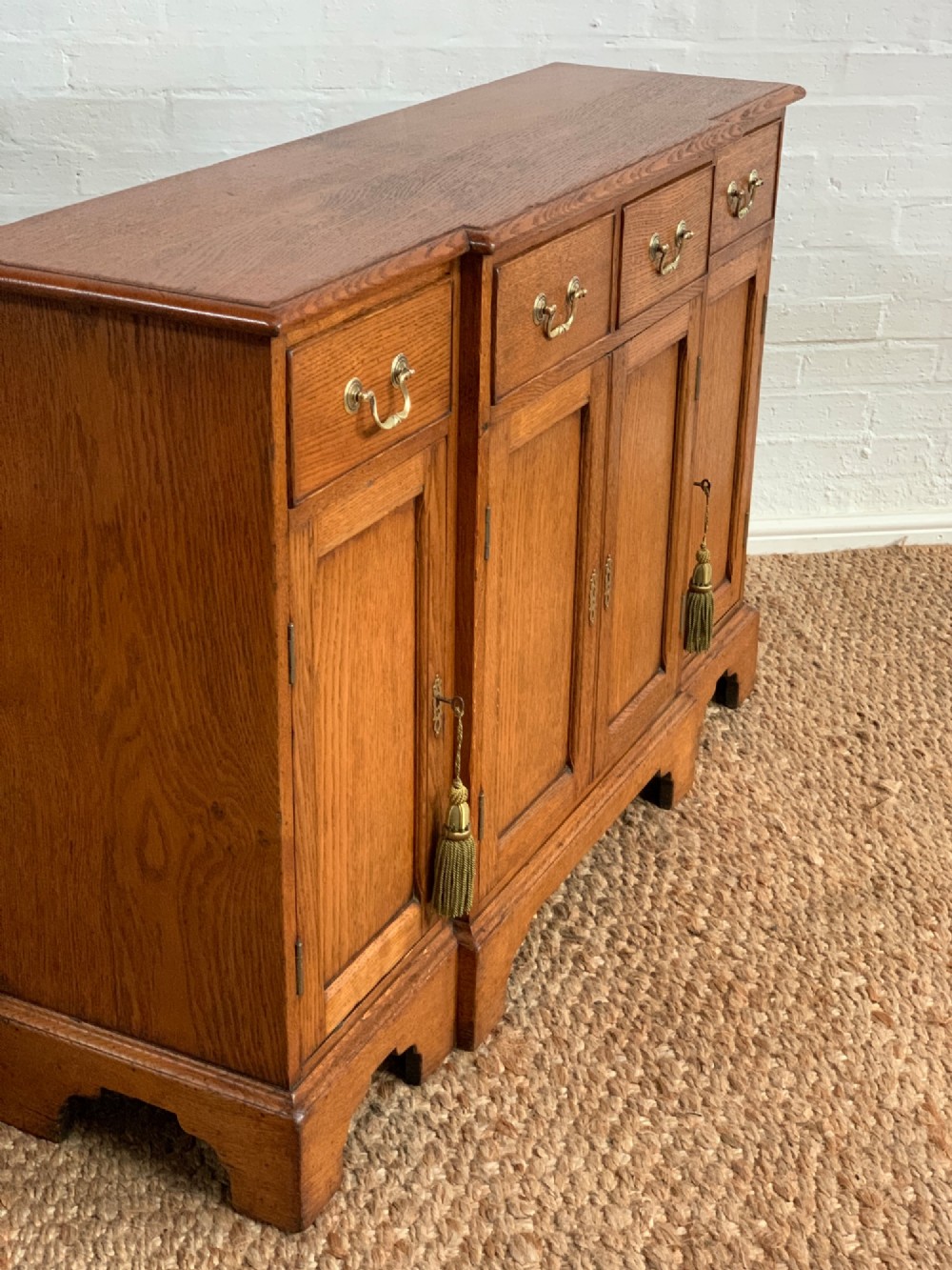 A golden oak sideboard featuring four upper drawers, four panelled cupboard doors with brass handles, keyhole escutcheons with tassels, and bracket feet. View IX
