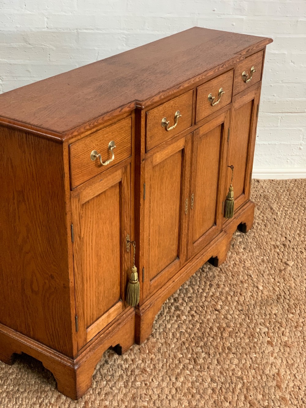 A golden oak sideboard featuring four upper drawers, four panelled cupboard doors with brass handles, keyhole escutcheons with tassels, and bracket feet. View X