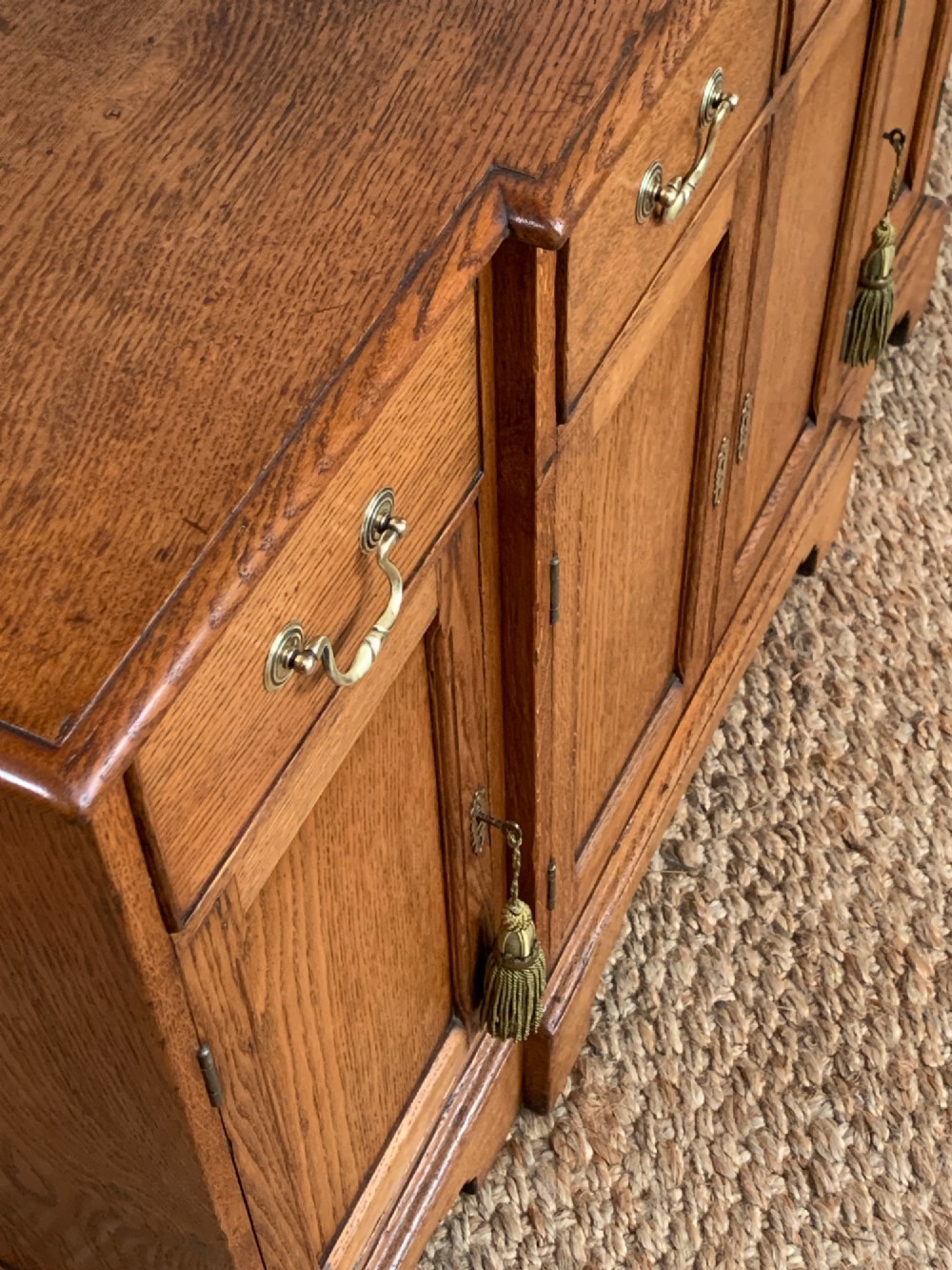 A golden oak sideboard featuring four upper drawers, four panelled cupboard doors with brass handles, keyhole escutcheons with tassels, and bracket feet. View XI