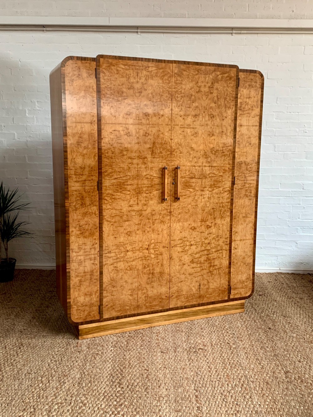An Art Deco double wardrobe crafted from figured burr walnut veneer with rounded top edges, vertical wooden handles, and a stepped plinth base.
