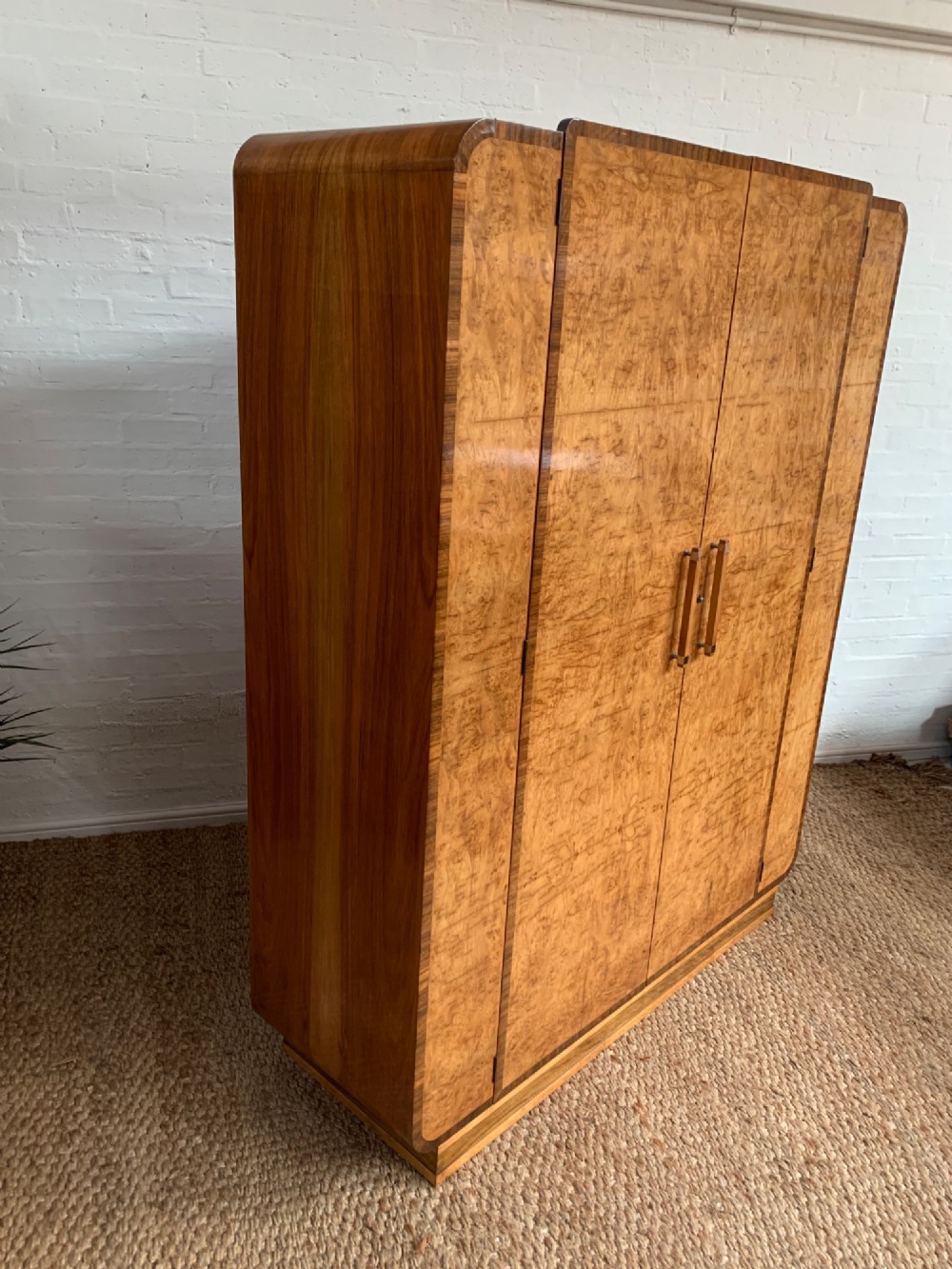 An Art Deco double wardrobe crafted from figured burr walnut veneer with rounded top edges, vertical wooden handles, and a stepped plinth base. View X