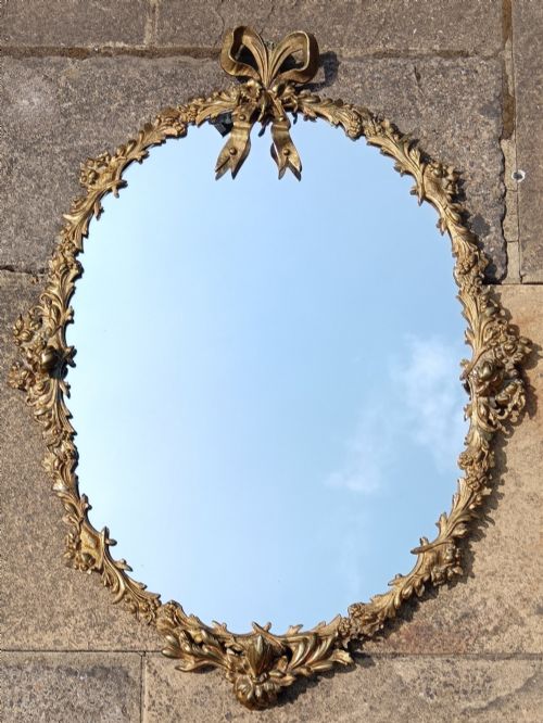 fig 1. An oval wall mirror framed in detailed gilt bronze with floral scrollwork and a prominent ribbon bow crest at the top.