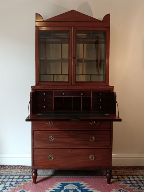 fig 2. A Regency mahogany secretaire cabinet featuring a triangular pediment top, glazed upper doors, a fall-front desk section, and three drawers below on turned feet.