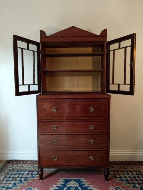 fig 3. A Regency mahogany secretaire cabinet featuring a triangular pediment top, glazed upper doors, a fall-front desk section, and three drawers below on turned feet.