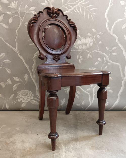 fig 1. A Victorian mahogany hall chair featuring a carved shield backrest with scroll detailing, a solid wooden seat, and turned front legs.