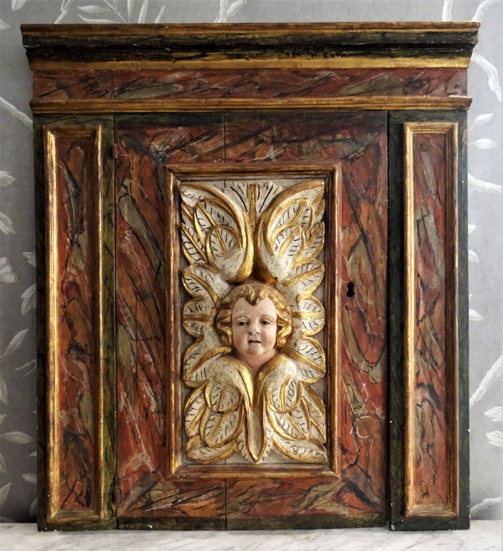 A rectangular Italian Baroque carved wood panel featuring a central gilded cherub head surrounded by leaf motifs, framed by faux-marbled painted sections and gilded mouldings.