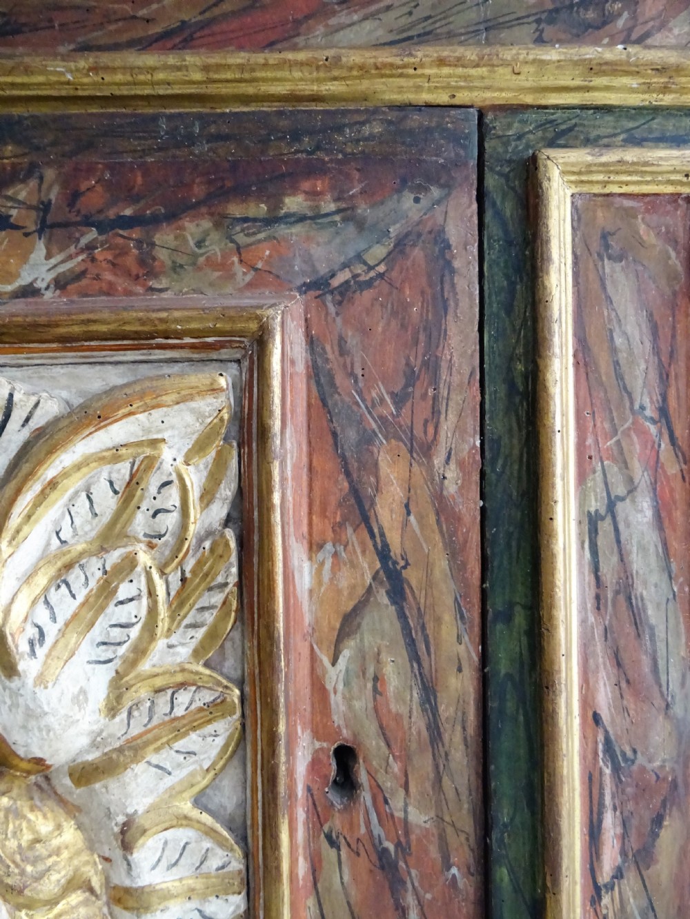 A rectangular Italian Baroque carved wood panel featuring a central gilded cherub head surrounded by leaf motifs, framed by faux-marbled painted sections and gilded mouldings. View II