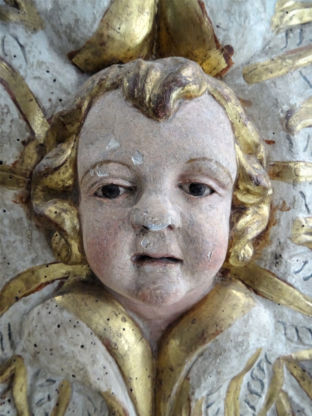 A rectangular Italian Baroque carved wood panel featuring a central gilded cherub head surrounded by leaf motifs, framed by faux-marbled painted sections and gilded mouldings. View V