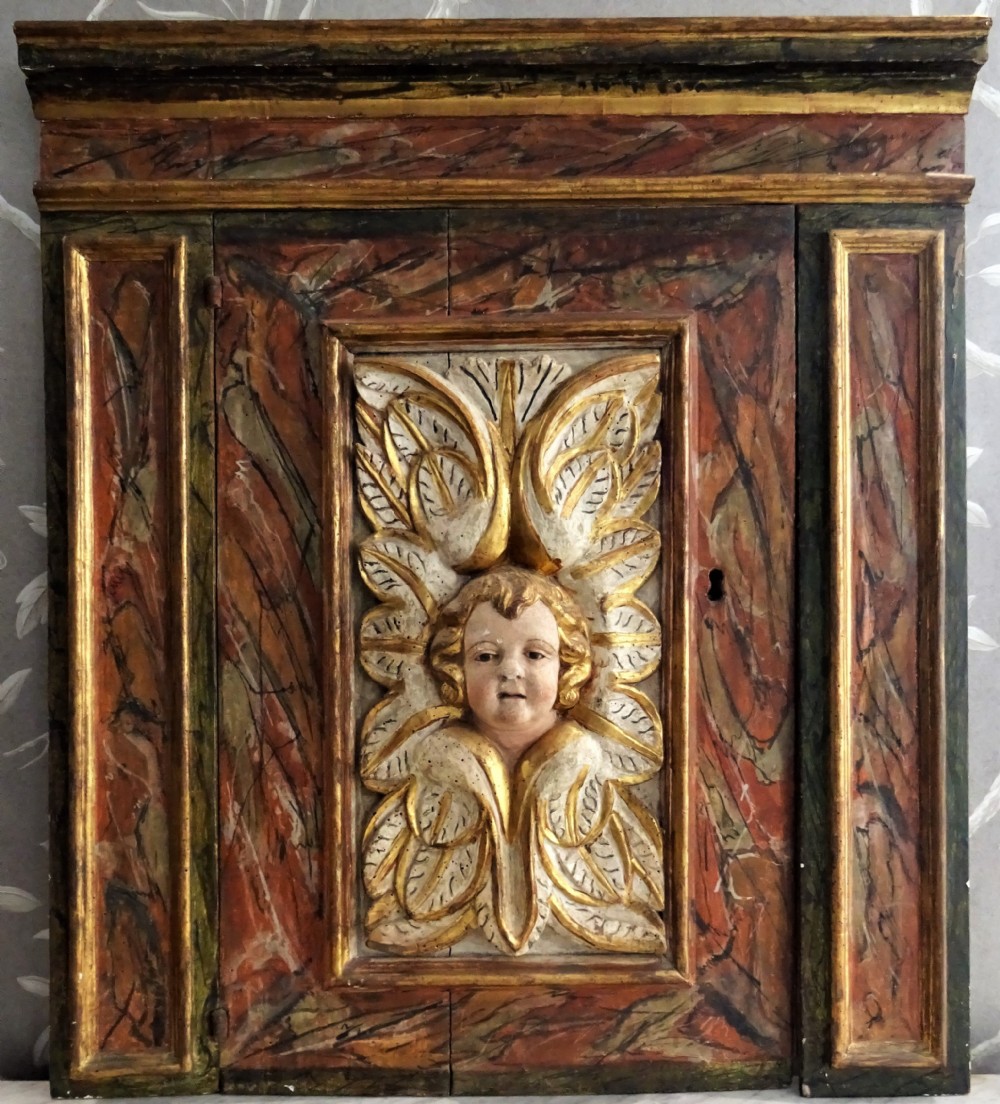 A rectangular Italian Baroque carved wood panel featuring a central gilded cherub head surrounded by leaf motifs, framed by faux-marbled painted sections and gilded mouldings. View VI