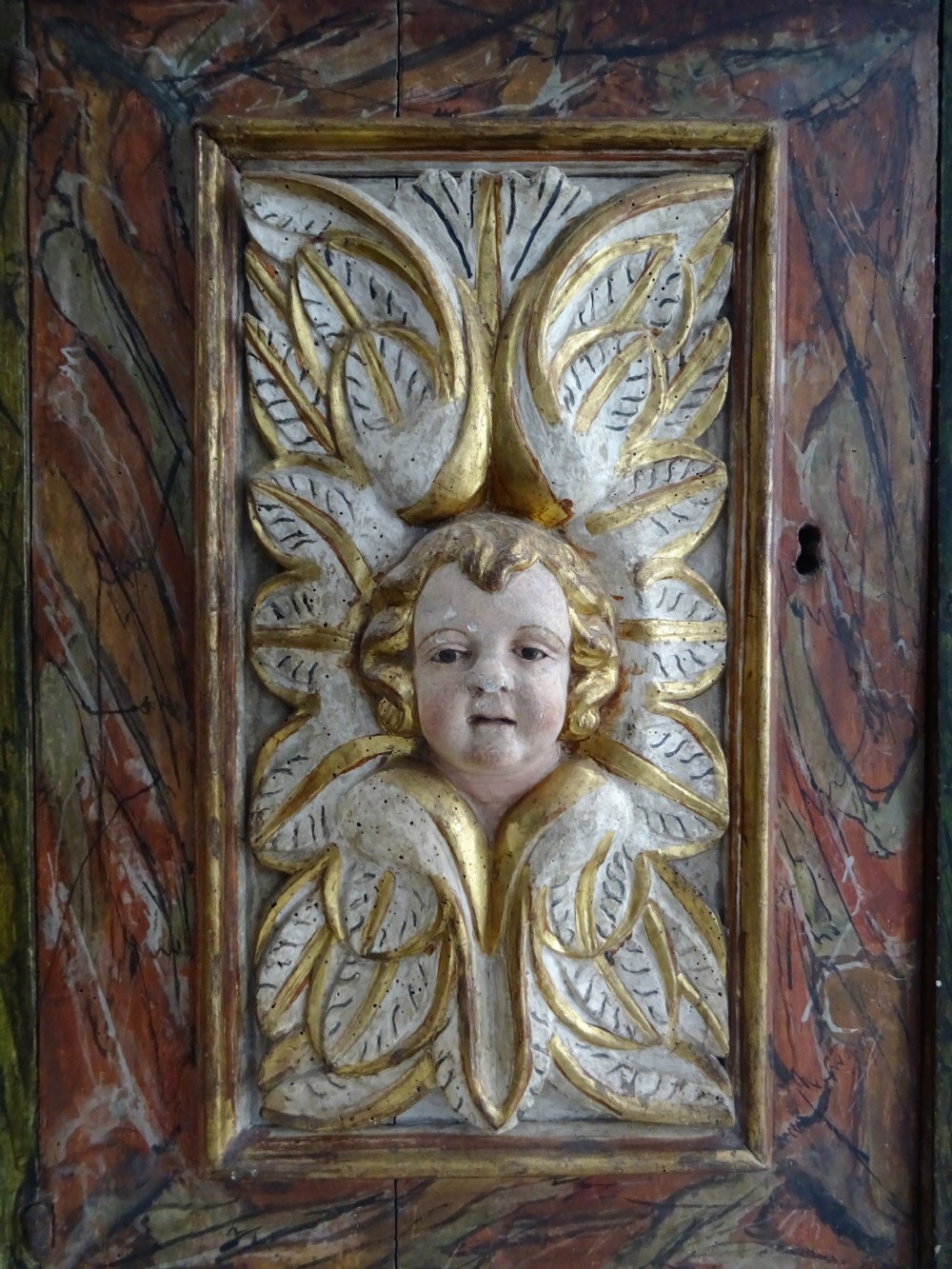 A rectangular Italian Baroque carved wood panel featuring a central gilded cherub head surrounded by leaf motifs, framed by faux-marbled painted sections and gilded mouldings. View IX