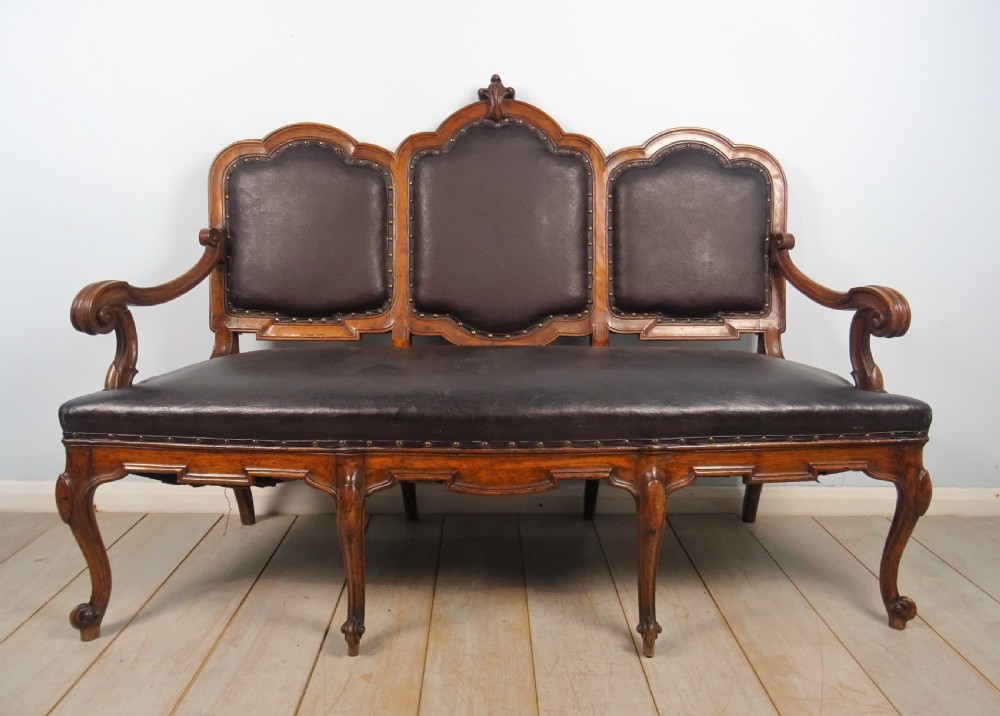 A 19th-century Italian settee featuring a carved walnut frame, three shaped back panels upholstered in dark brown leather with brass studs, scrolled arms, and cabriole legs.