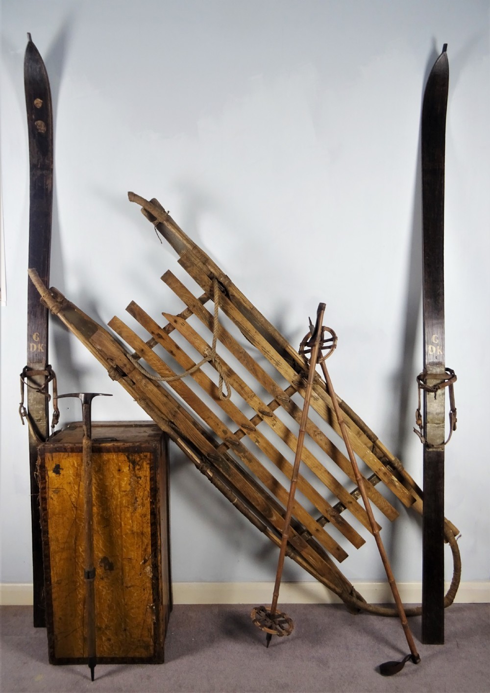 A collection of antique expedition equipment including tall wooden skis with leather bindings, bamboo ski poles, an ice axe, a wooden slatted sledge, and a wooden chest.