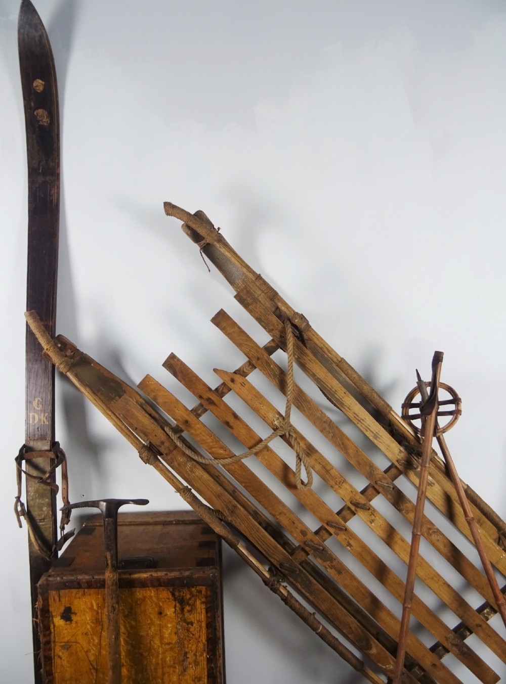 A collection of antique expedition equipment including tall wooden skis with leather bindings, bamboo ski poles, an ice axe, a wooden slatted sledge, and a wooden chest. View II