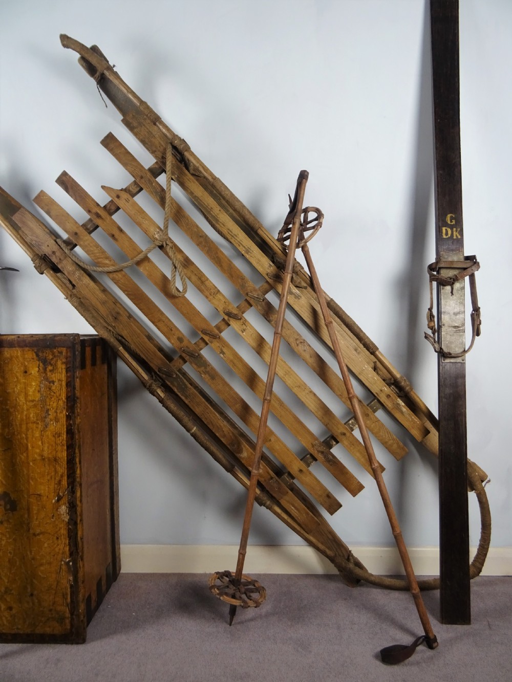 A collection of antique expedition equipment including tall wooden skis with leather bindings, bamboo ski poles, an ice axe, a wooden slatted sledge, and a wooden chest. View III